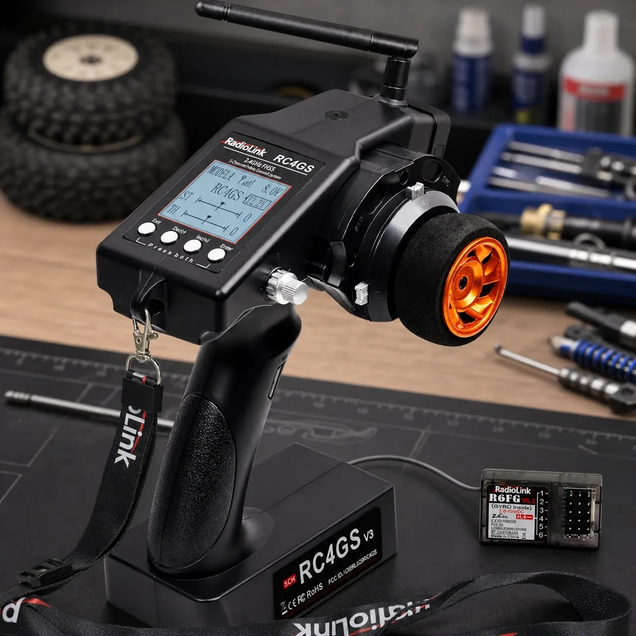Close-up of a Radiolink RC4GS V3 surface RC transmitter on a workshop bench, with orange steering wheel accent, antenna extended, lanyard attached, and receiver module shown beside it amid hobby tools and parts.